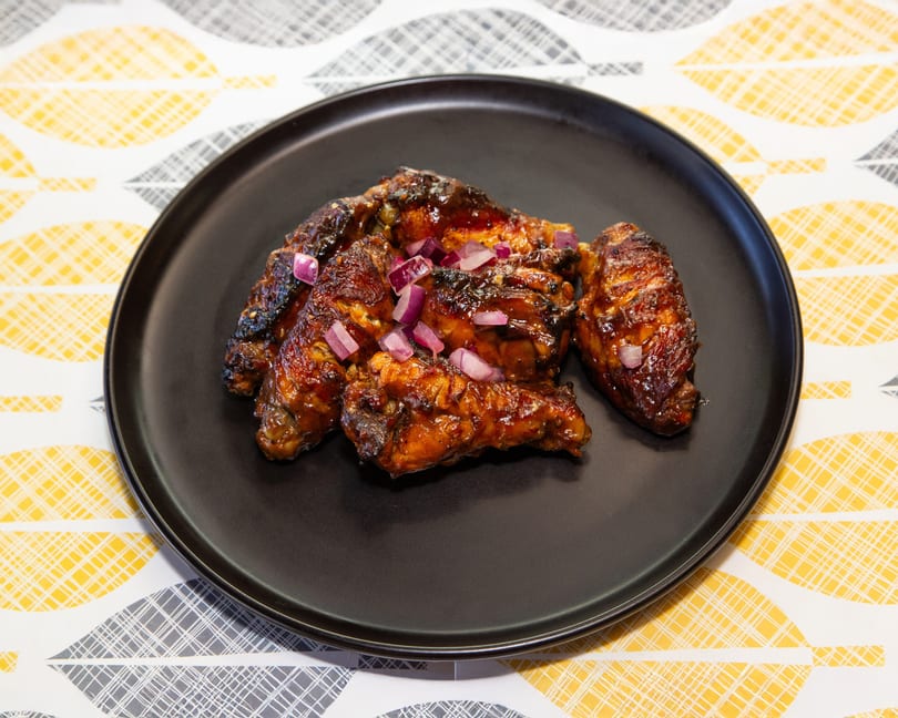 Glazed chicken wings topped with diced red onions on a black plate, on a patterned tablecloth.