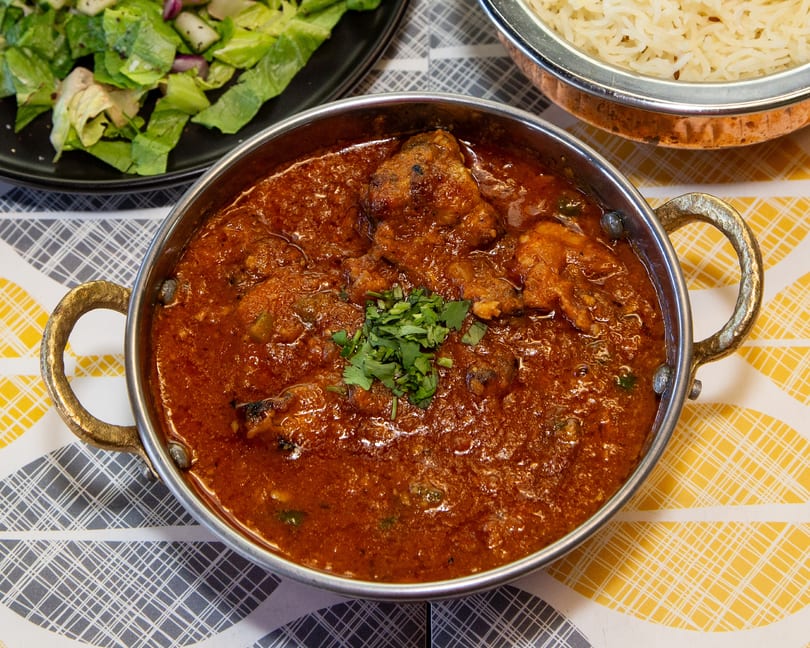 Chicken curry in a metal bowl, garnished with cilantro, alongside rice and salad.